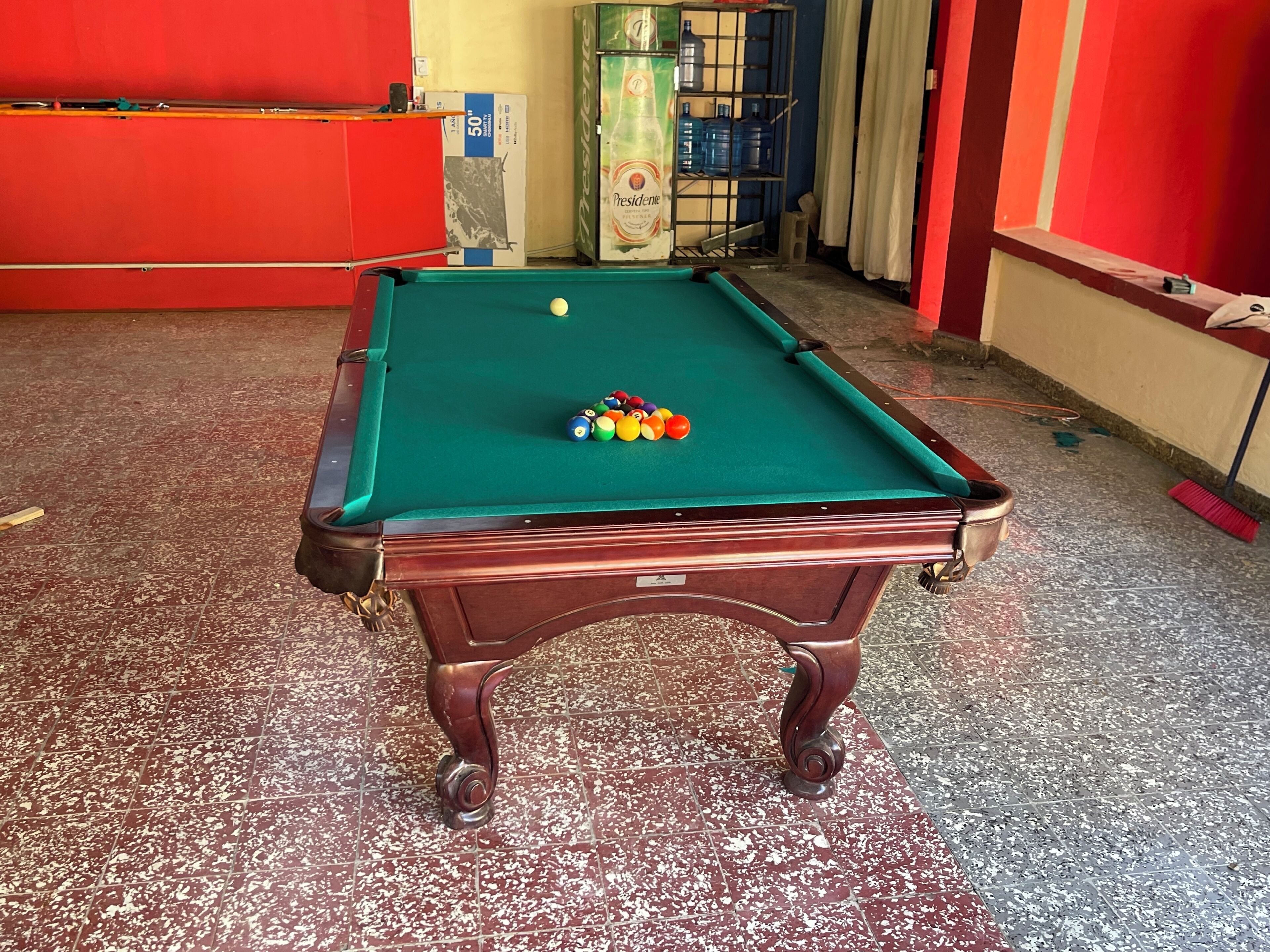 Une table de billard avec des billes et une queue, entourée d’un mur rouge et posée sur un sol carrelé.