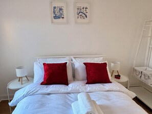 A neatly made bed with white bedding and two red pillows, a bedside table with a lamp, and two framed cityscape pictures on the wall. - Spacious Victorian House - E6 London (London)