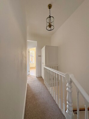 A hallway with a carpeted floor, white walls, a white railing, and a hanging light fixture. - Spacious Victorian House - E6 London (London)