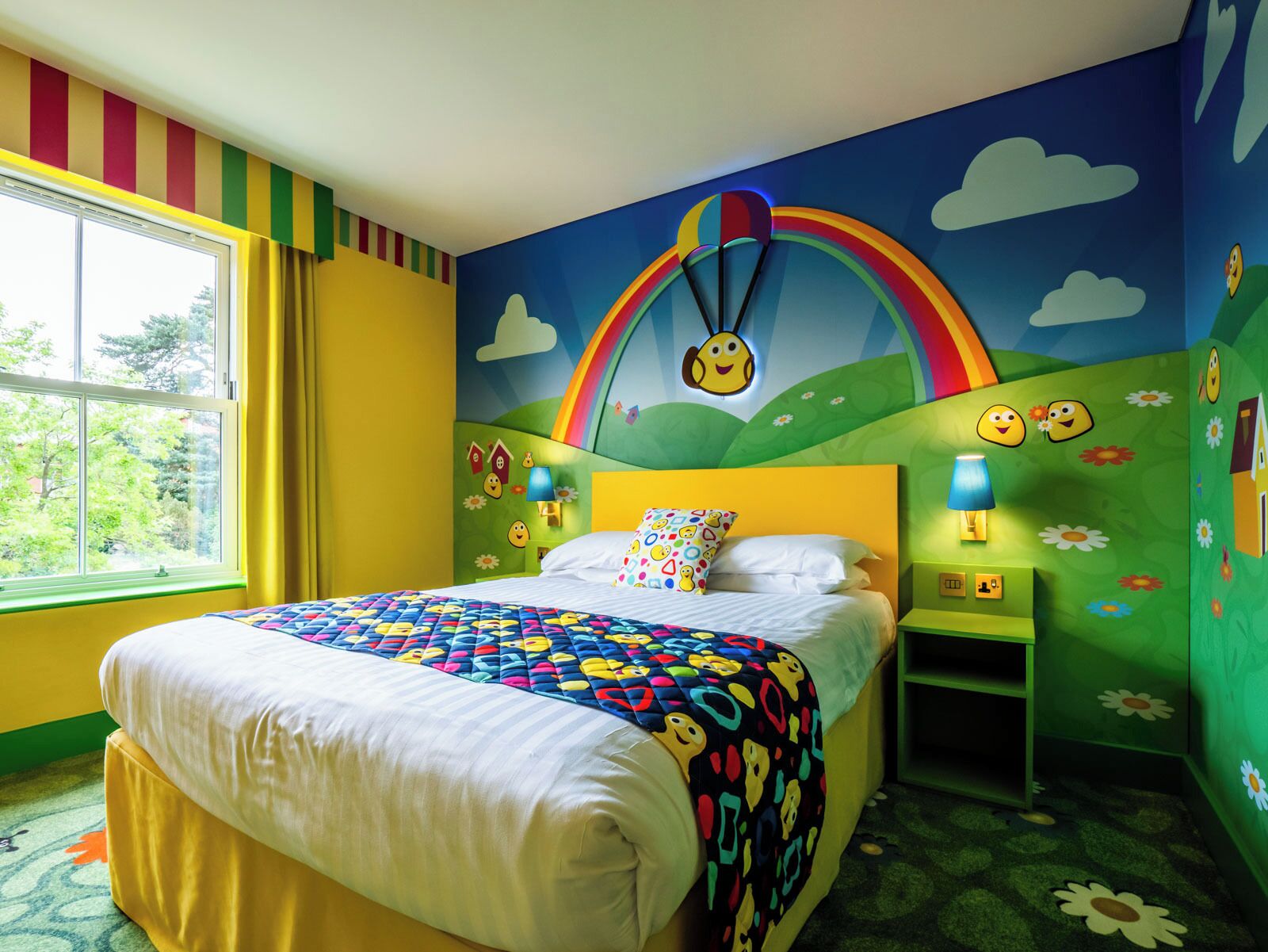 Habitación infantil con cama, ventana y un mural que muestra un arcoíris y caras sonrientes.