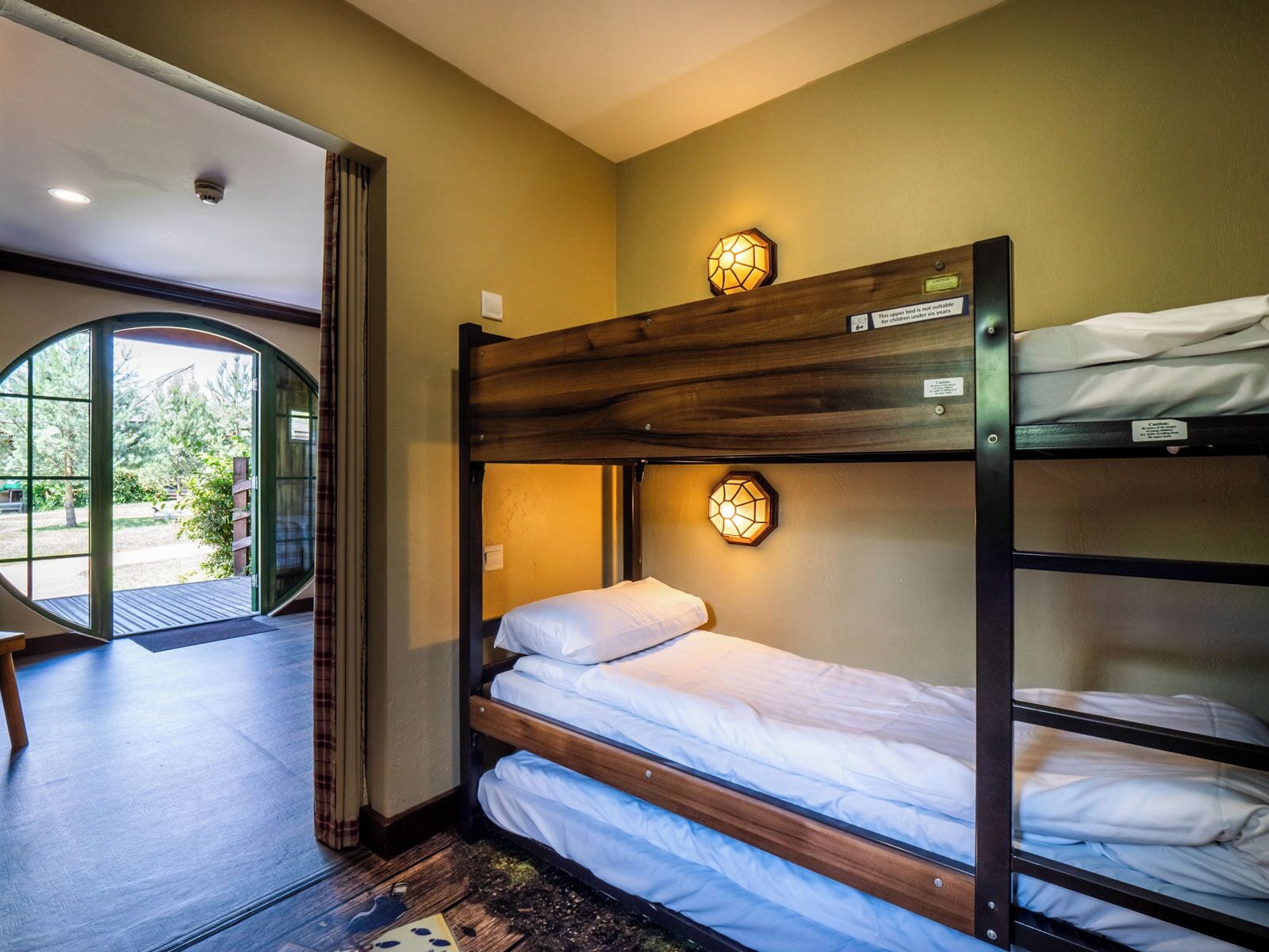 Une chambre avec un lit superposé, une tête de lit en bois, une petite fenêtre et une porte donnant sur l’extérieur.