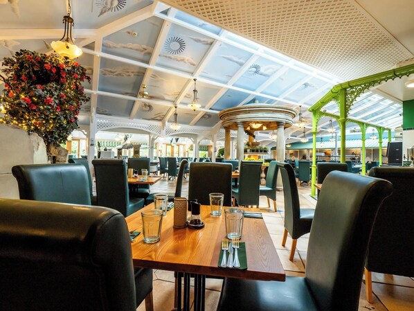 A spacious restaurant with high ceilings, a central dining area with tables and chairs, and a decorative hanging plant.