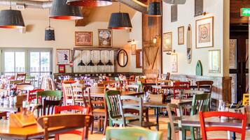 Un interior de restaurante con varias mesas y sillas, luces colgantes y diferentes cuadros en las paredes.