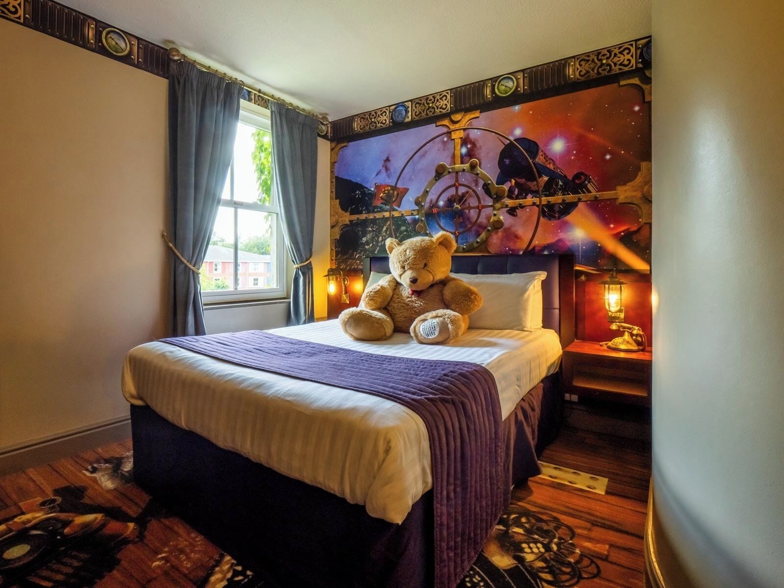 A hotel room with a large bed, a teddy bear on the bed, a window with curtains, a wall mural, and a nightstand with a lamp.