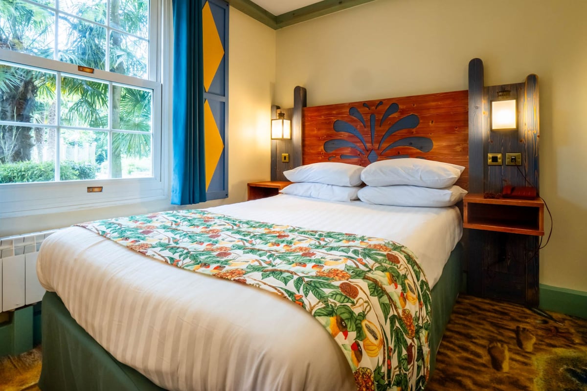 Quarto de hotel com cama, janela com cortinas, cabeceira com design decorativo, luminárias de cabeceira e mesa de cabeceira de madeira.
