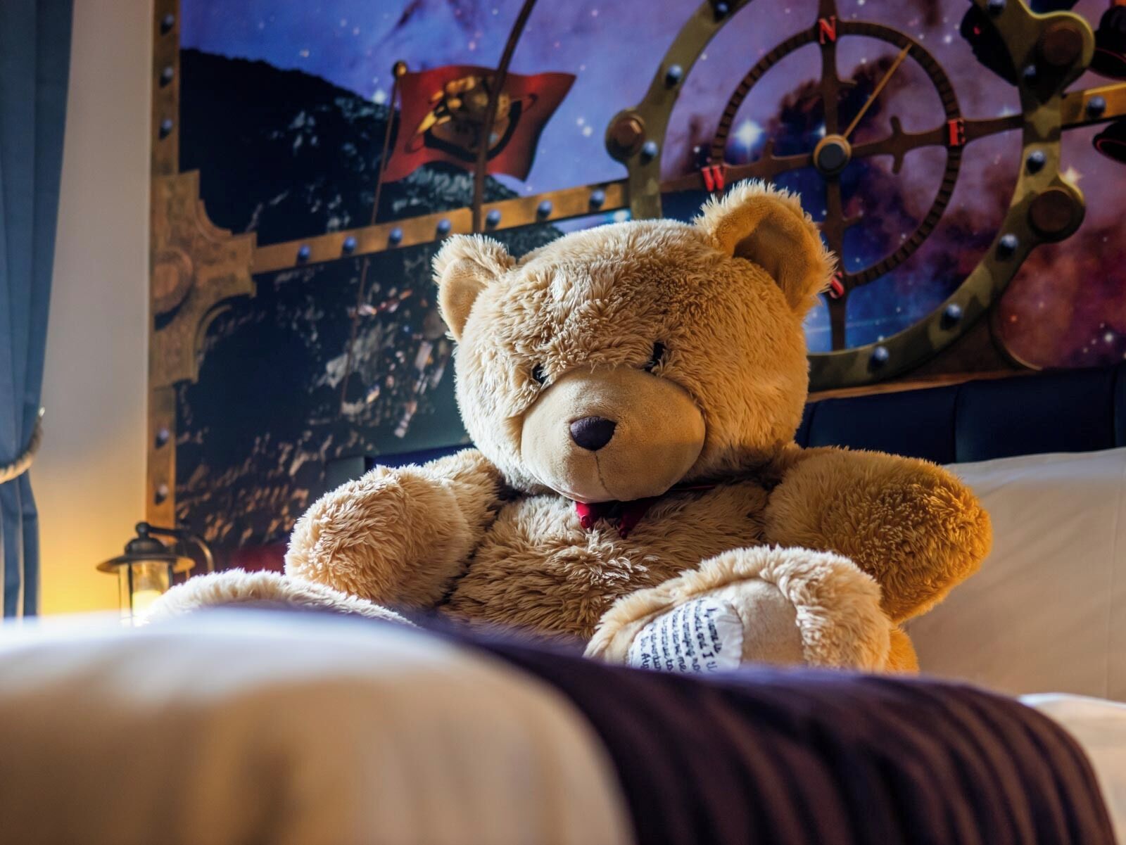A plush teddy bear on a bed with a starry night backdrop and a ship's wheel.
