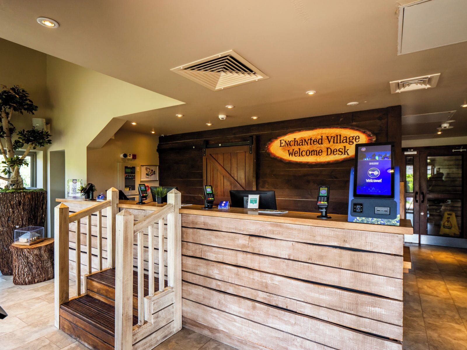 A reception desk with a wooden finish, a digital kiosk, and a sign reading 'Enchanted Village Welcome Desk.'