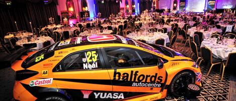 A luxury hotel banquet hall with round tables set for dining, a luxury car parked in the foreground, and a stage with a screen in the background.