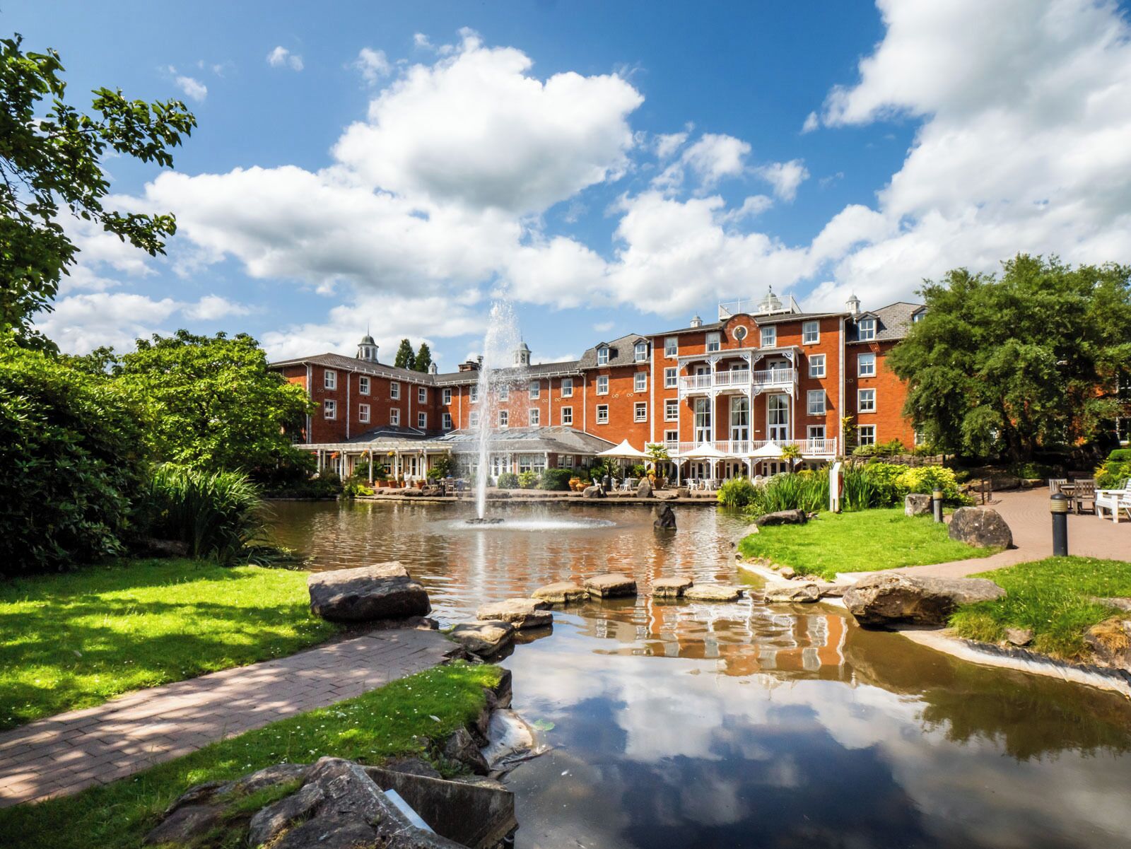 A large red brick building with a fountain in the center, surrounded by a well-maintained garden and a pond.