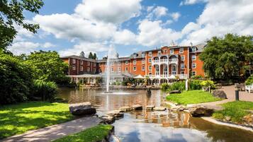 A large red brick building with a fountain in the center, surrounded by a well-maintained garden and a pond.