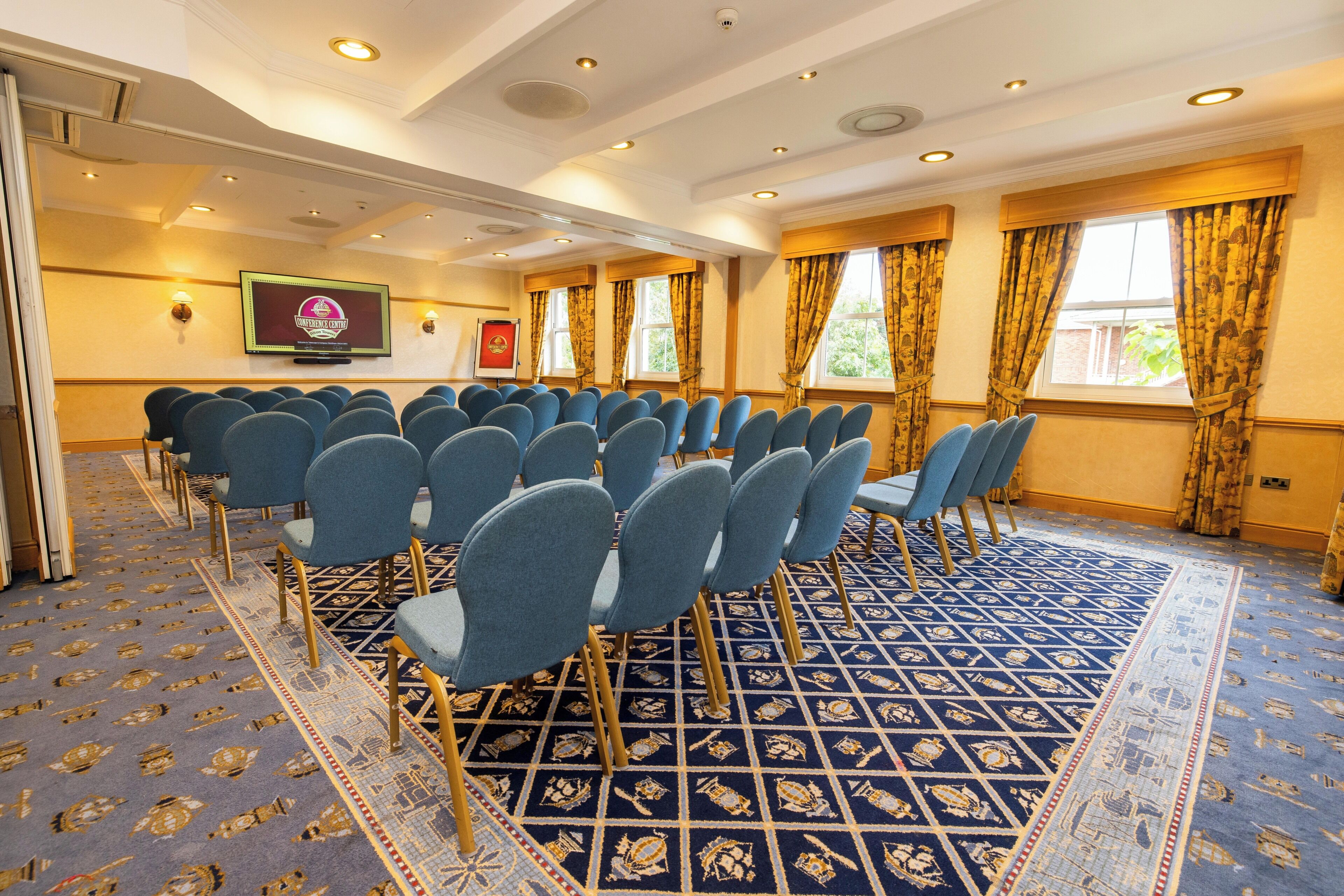A conference room with rows of chairs, a carpeted floor, and a screen displaying a logo.
