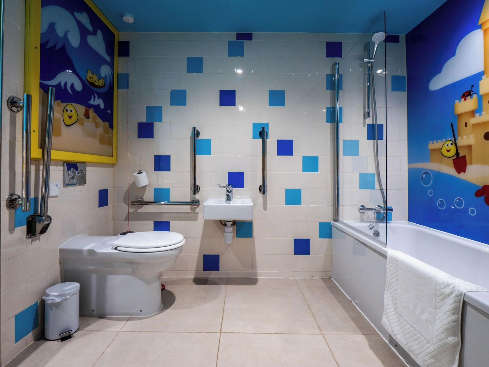 A bathroom with a toilet, sink, and bathtub, featuring blue square tiles and cartoonish wall art.