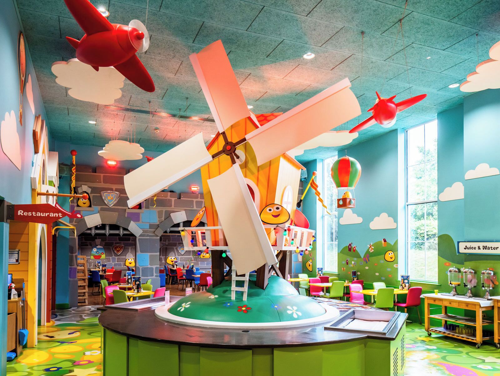 A children's play area with a large windmill structure, tables, chairs, and a juice and water station.
