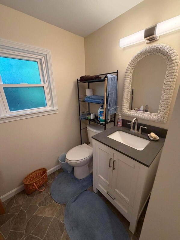 A bathroom with a toilet, sink, and a mirror. There is a window above the toilet and a basket next to it.