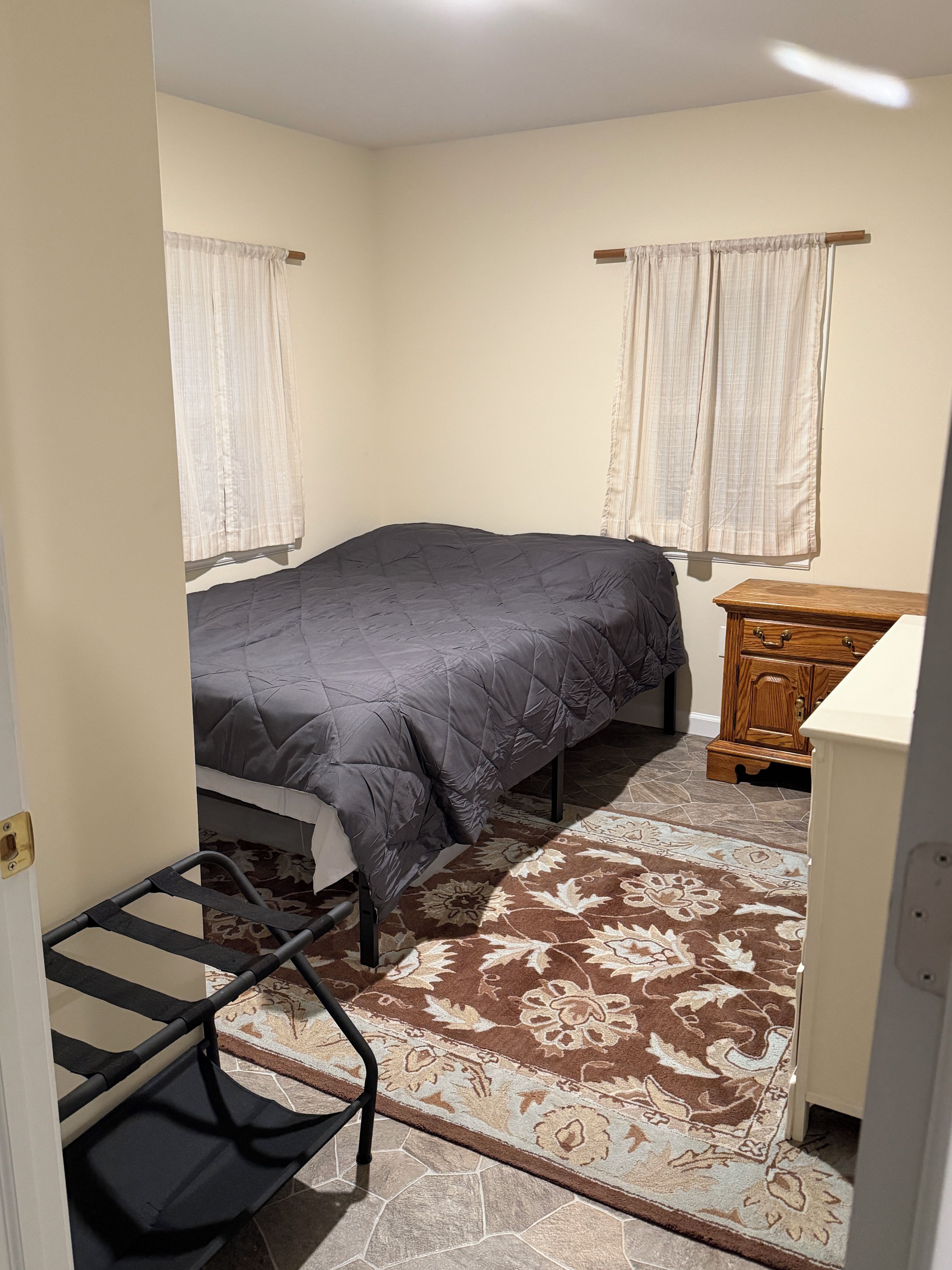 A bedroom with a bed, a wooden dresser, a patterned rug, and a window with curtains.