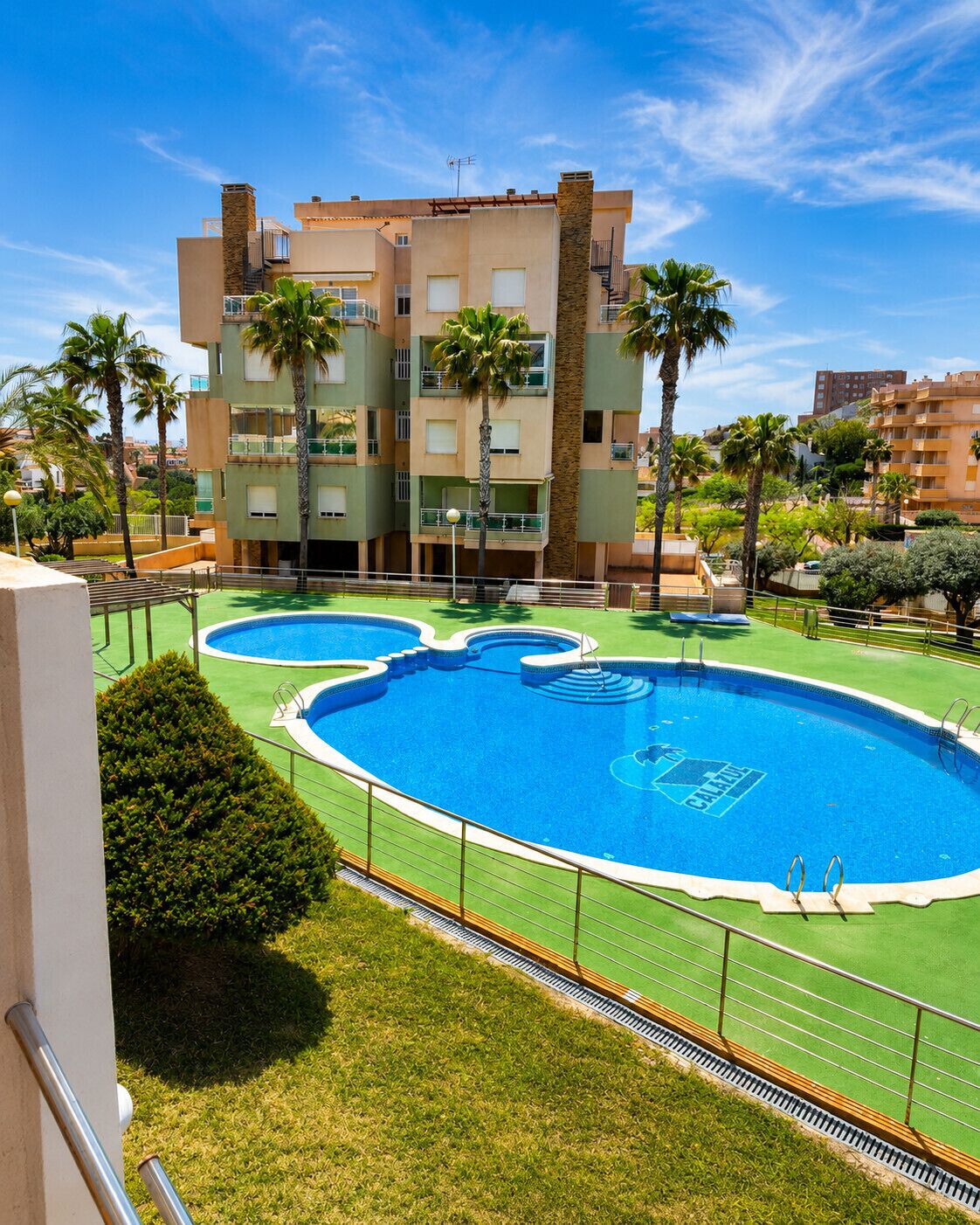 A multi-story building with a swimming pool, palm trees, and a clear sky.