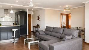 A modern living room with a grey sectional sofa, a glass coffee table, and a kitchen area with a stainless steel refrigerator. - Cala Flores Residence – 3-Bedroom, Pool & Terrace (Cabo de Palos)