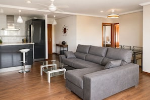 A modern living room with a grey sectional sofa, a glass coffee table, and a kitchen area with a stainless steel refrigerator. - Cala Flores Residence – 3-Bedroom, Pool & Terrace (Cabo de Palos)