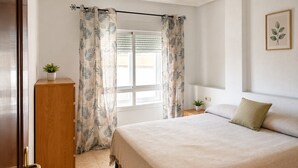 A bedroom with a bed, a wooden wardrobe, a ceiling fan, a window with curtains, and a small bedside table with a plant. - Cala Flores Residence – 3-Bedroom, Pool & Terrace (Cabo de Palos)