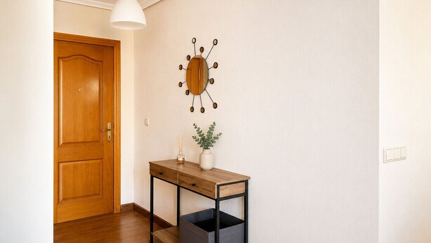 A hallway with a wooden door, a wall-mounted decorative clock, a small table with a vase and plant, and a trash bin. - Cala Flores Residence – 3-Bedroom, Pool & Terrace (Cabo de Palos)