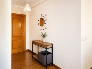 Un corridoio con una porta in legno, un orologio decorativo a parete, un tavolino con un vaso e una pianta, e un cestino.