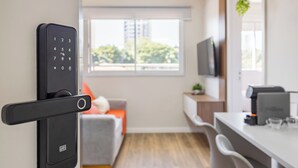 A modern room with a door lock, a sofa with a cushion, a dining area with chairs, a television, and a window with a view of buildings. - StayHome Chácara Klabin - AC & Gym (São Paulo)