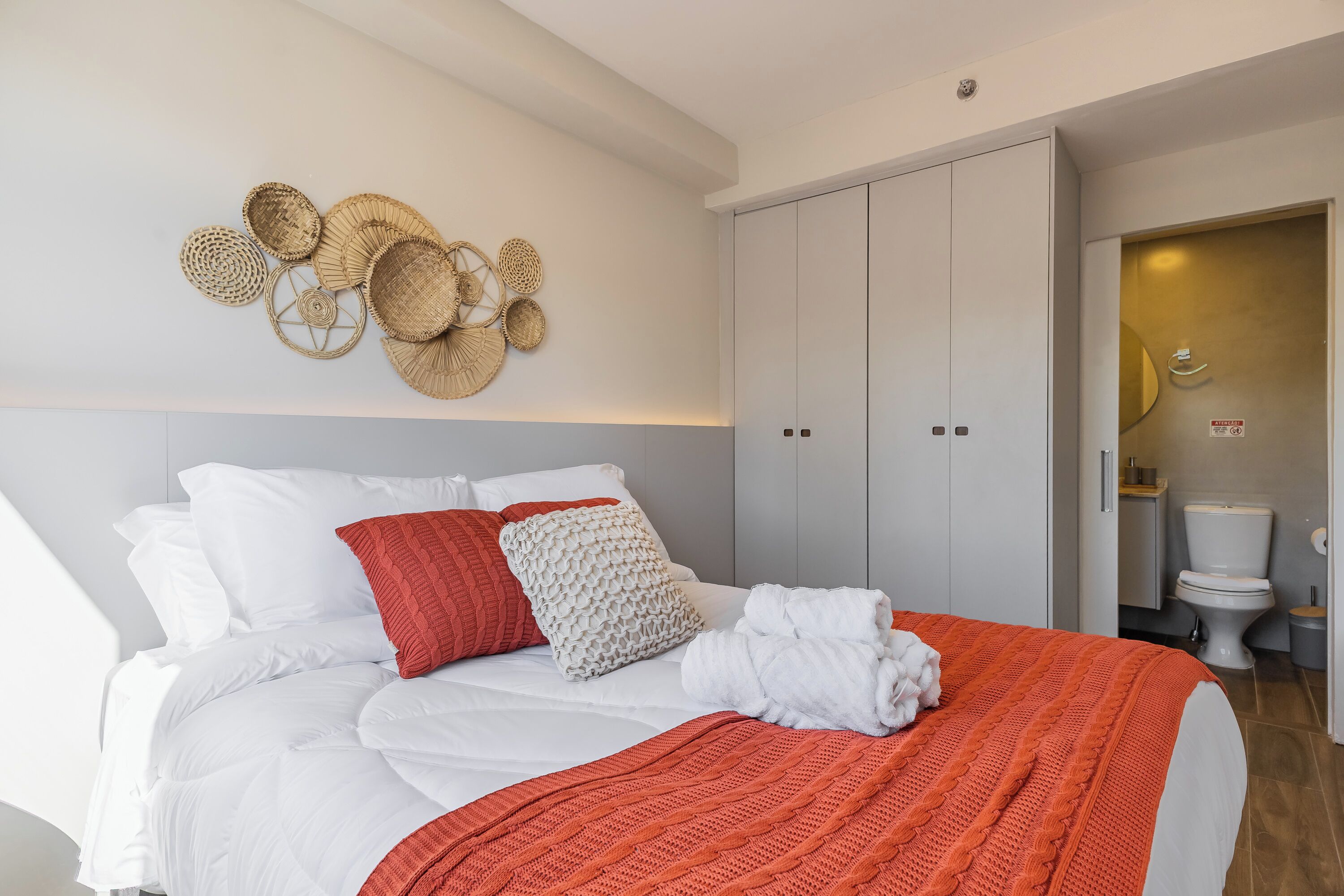 A neatly made bed with white and red bedding, a wall-mounted woven art piece, and a closed wardrobe.