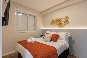 Una cama bien hecha con edredón blanco y detalles naranja, una ventana grande con persianas, y un objeto decorativo colgado en la pared.