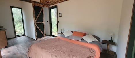 Un dormitorio con cama, almohadas, una mesita de noche y vistas a través de una puerta abierta.