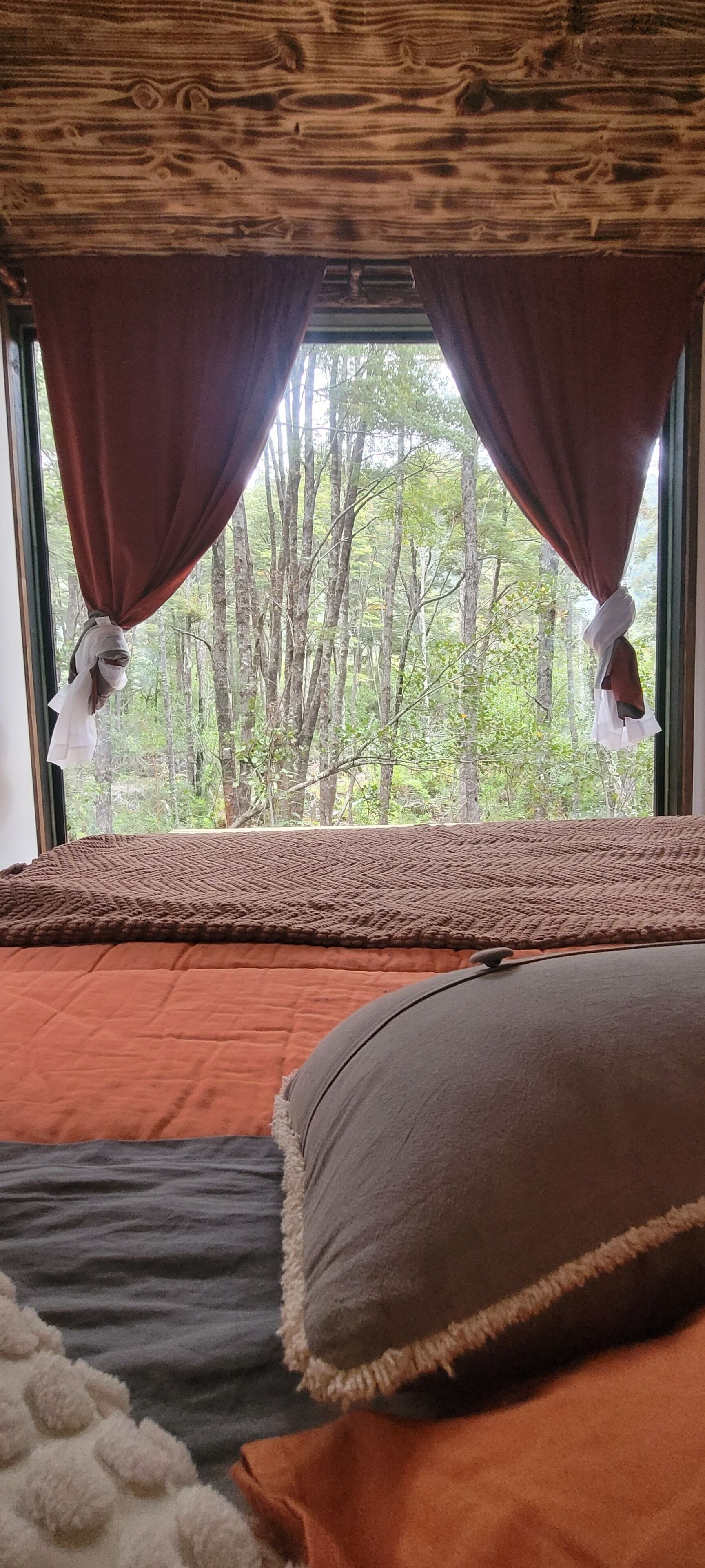 A bedroom with a large window, a bed with an orange bedspread, a brown pillow, and a view of trees outside.