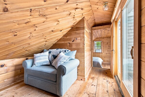 A cozy attic bedroom with a wooden ceiling, a gray sofa with pillows, and a window with blinds.