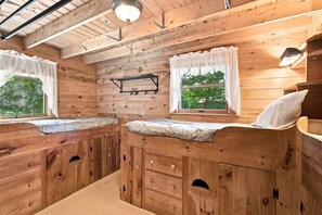 Interior de una cabaña de madera con dos camas, una estantería y una ventana con cortinas.