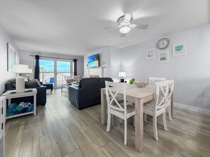 A modern living room with a dining area, a ceiling fan, and a TV mounted above a fireplace. - Ocean-View Oasis: 2BR Condo w/ Pool & Private Balcony (Indian Shores)