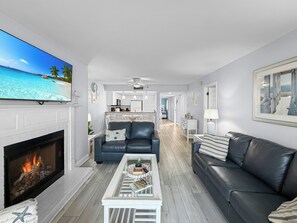 A living room with a fireplace, a sofa, a coffee table, and a television displaying a beach scene. - Ocean-View Oasis: 2BR Condo w/ Pool & Private Balcony (Indian Shores)