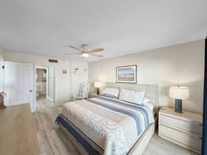 Un dormitorio con cama, mesitas de noche, ventilador de techo, un cuadro en la pared y una puerta que da a un baño.