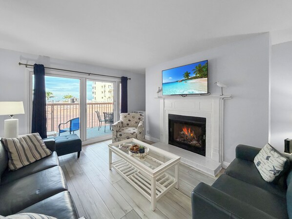 Una sala de estar con chimenea, televisor, sofá, mesa de centro y balcón con vista al mar.