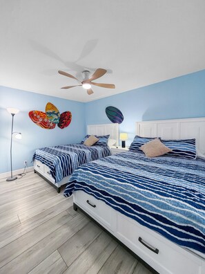 A bedroom with two beds, a ceiling fan, and wall art. - Ocean-View Oasis: 2BR Condo w/ Pool & Private Balcony (Indian Shores)