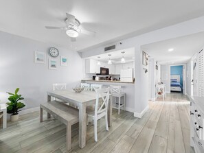 A modern kitchen with white cabinetry, a dining area with a wooden table and chairs, and a hallway leading to a bedroom. - Ocean-View Oasis: 2BR Condo w/ Pool & Private Balcony (Indian Shores)