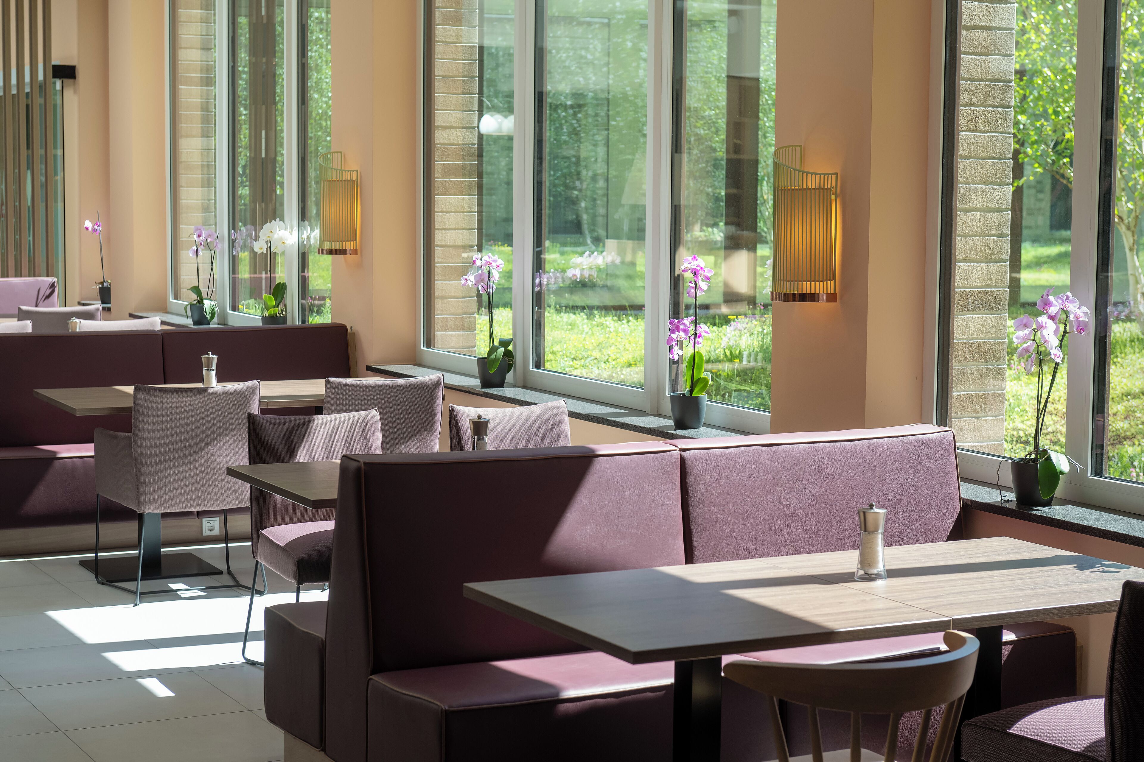 A modern indoor seating area with tables and cushioned benches, large windows with a view of greenery, and potted plants.