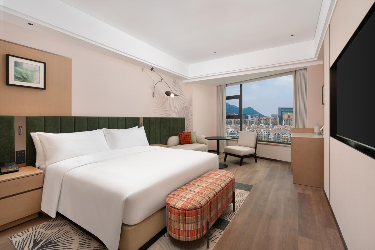 Ein Hotelzimmer mit einem großen Bett, einem Schreibtisch, einem Sessel und Blick auf die Stadt.
