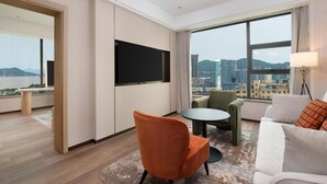 A modern hotel room with a large window, a flat-screen TV, a sofa, a chair, and a small table. - Hilton Garden Inn Zhoushan Putuo District (Zhoushan)
