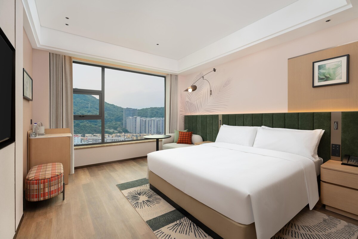 Ein modernes Hotelzimmer mit einem großen Bett, Blick auf die Stadt und einer kleinen Sitzecke.