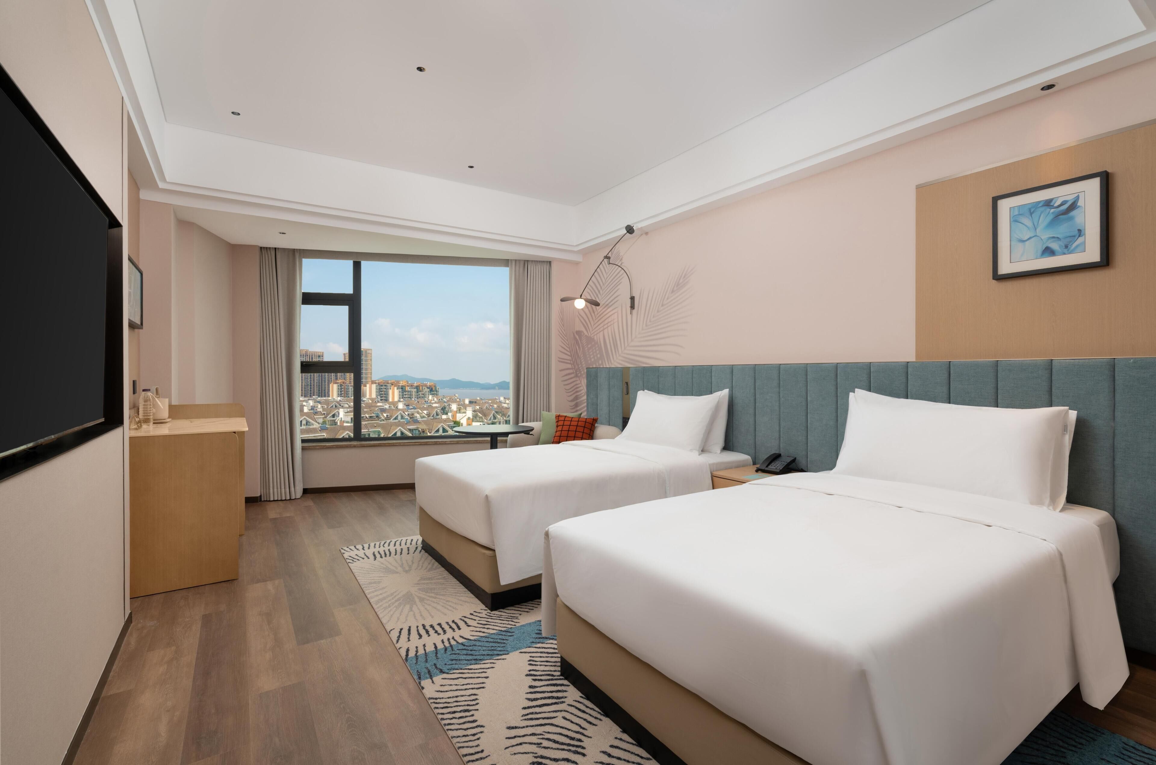 Habitación de hotel con dos camas, un escritorio, una silla, una ventana con vista a la ciudad y una obra de arte colgada en la pared.