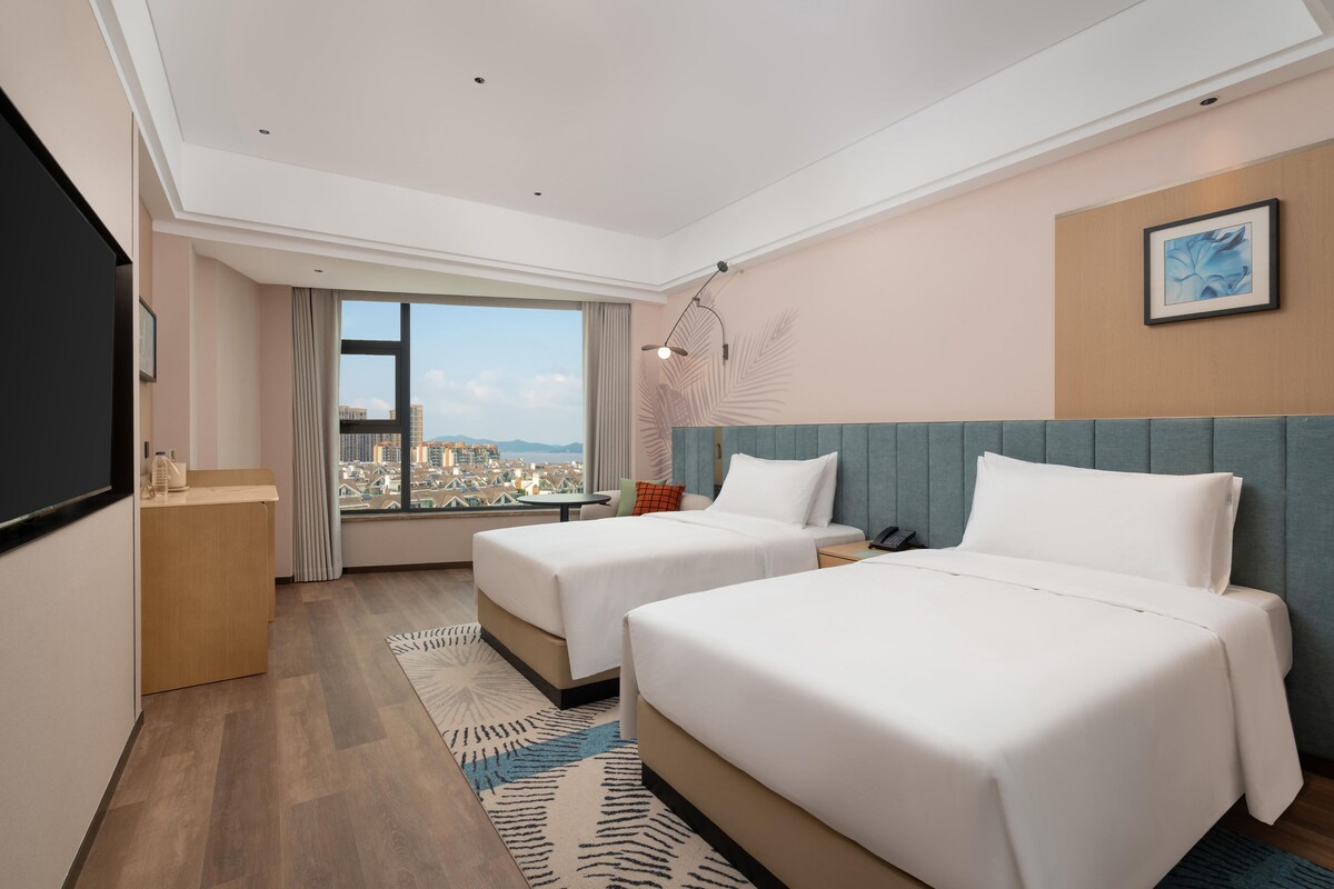 Ein Hotelzimmer mit zwei Betten, einem Schreibtisch, einem Stuhl, einem Fenster mit Stadtblick und einem Wandbild.
