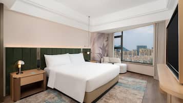 A modern hotel room with a large bed, a sitting area, a TV, and a city view.