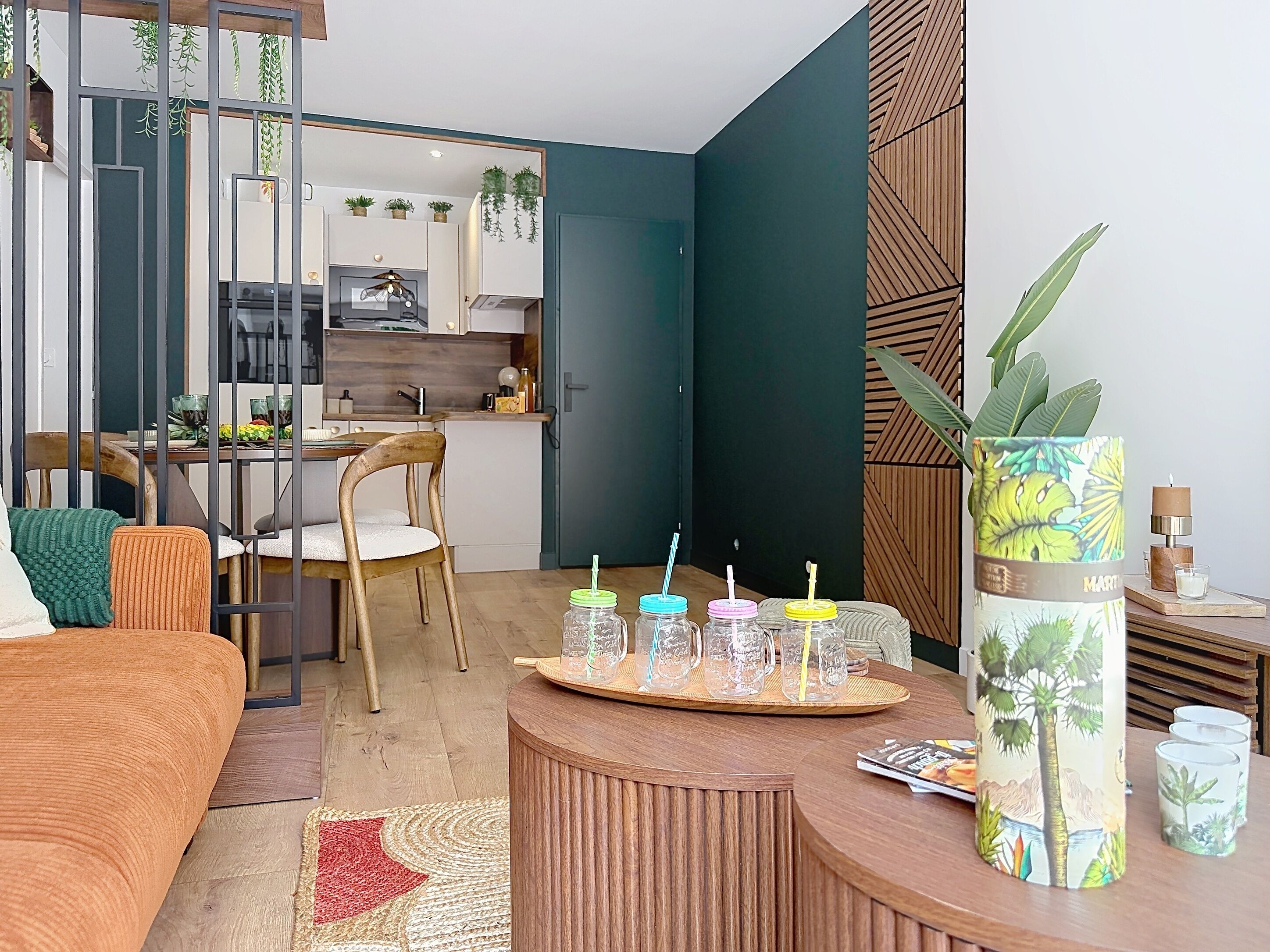 A modern living room with a wooden sofa, a dining table with chairs, a kitchen in the background, and a decorative plant on a side table.