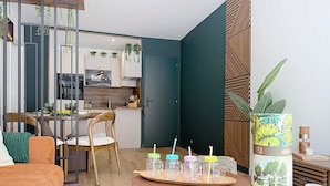 Un salon moderne avec un canapé en bois, une table à manger avec des chaises, une cuisine en arrière-plan et une plante décorative posée sur une table d’appoint.
