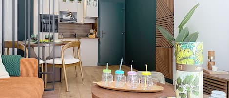 Een moderne woonkamer met een houten bank, een eettafel met stoelen, een keuken op de achtergrond en een decoratieve plant op een bijzettafeltje.