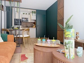 A modern living room with a wooden sofa, a dining table with chairs, a kitchen in the background, and a decorative plant on a side table. - Love of the Islands • Center • Beach • Parking (Cabourg)