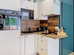A modern kitchen with white cabinetry, a wooden accent wall, a built-in microwave, a coffee maker, and a small countertop with a box of biscuits. - Love of the Islands • Center • Beach • Parking (Cabourg)